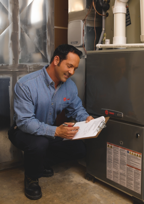 A technician in a blue shirt kneels by an HVAC unit, holding a clipboard and pen, performing HVAC maintenance as he inspects and records vital system information.